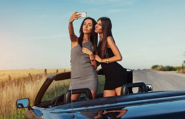 Two sexy brunette woman standing near cabriolet on the road in pitch and pictures to your phone.