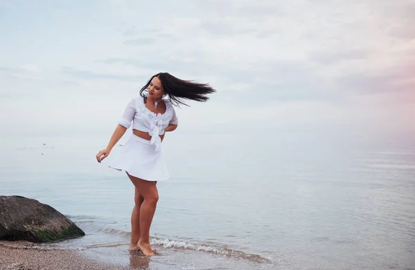 woman in a white shirt and skirt dancing barefoot on the sand beach hand above the azure sea breeze shakes her long hair