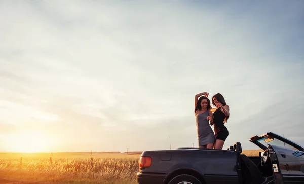 Young two women at a photo shoot. Girls gladly posing next to a black car against the sky on a fantastic sunset.