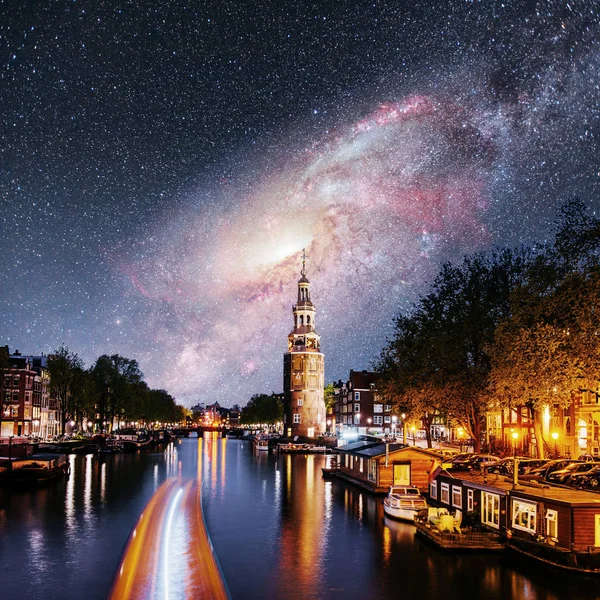 Beautiful night in Amsterdam. Night illumination of buildings and boats near the water in the channel. Fantastic starry sky and the milky way. Courtesy of NASA.