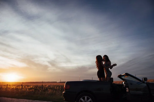 Young two women at a photo shoot. Girls gladly posing next to a black car against the sky on a fantastic sunset.