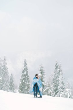 Genç yakışıklı adam onun genç ve güzel kız hugs ve onu yün chekered mavi battaniye ile kapsar. Snowy orman kış yürüyüş. Kışın seviyorum.