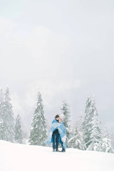 年轻英俊的男人拥抱他漂亮的年轻女孩 用羊毛的面颊蓝色毯子覆盖她 冬天在雪林中散步 冬天的爱 — 图库照片