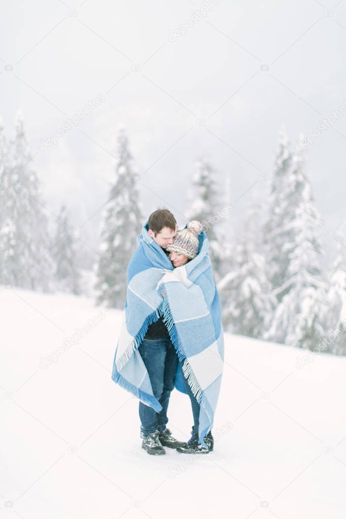 Young handsome man hugs his pretty young girl and covers her with the wool chekered blue blanket. Winter walk in snowy forest. Love in winter