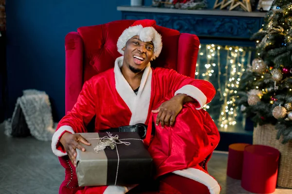 クリスマスと新年あけましておめでとうございますのコンセプトです アフリカ男性サンタ クロース クリスマス プレゼントとギフト ボックス赤袋を押しながらクリスマス ツリー近くの赤い椅子に座っての笑顔 — ストック写真