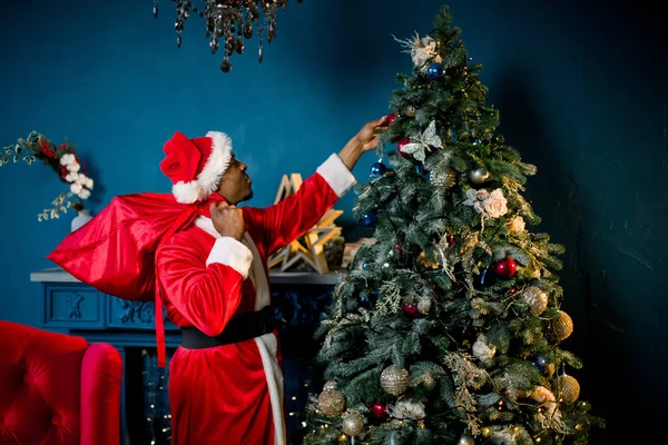 アフリカのサンタ クロースは クリスマス ツリーのそばに立ちます 笑って 笑ってアフリカ男性サンタ クロース居心地の良い青と赤の装飾が施された部屋でクリスマス プレゼントで大きな布の袋を保持しています — ストック写真