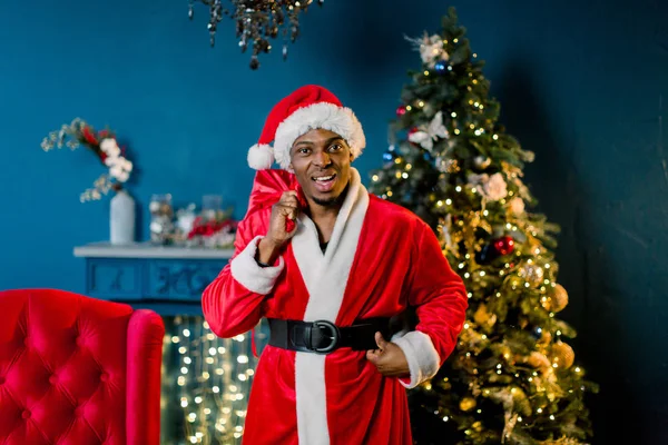 サンタ クロースはクリスマスの準備ができて 笑って 笑ってアフリカ男性サンタ クロースの飾りが付いている部屋に立っている間クリスマスのギフト袋を保持しています — ストック写真