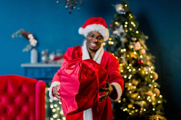 居心地の良い装飾が施された部屋クリスマス ツリーの前に立っているとプレゼントが付いている赤い袋を保持している彼の赤い衣装で幸せなアフリカ サンタ — ストック写真