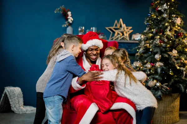 Kinderen Knuffelen Afrikaanse Santa Claus Zittend Een Rode Stoel Achtergrond — Stockfoto