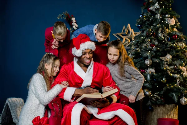 サンタ クロースが赤い椅子の周りに座って子供たちのクリスマス話で古代の本を読み取ります — ストック写真