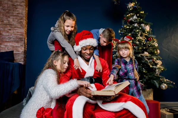 小さな美しい子供がサンタ クロースがクリスマス ツリーの背景の赤い椅子に座って読むの話に注意深く耳を傾ける — ストック写真
