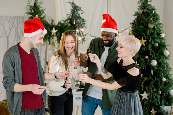 Glad men and satisfied ladies drinking glasses of champagne while communicating and laughing on New Year party. They keeping indian lights in hands during party