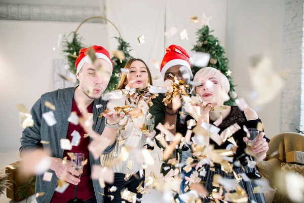 クリスマス パーティーで笑いながら友人のグループです 大晦日のパーティーで深夜黄金紙ふぶきを吹いて サンタさんの帽子を身に着けている つの美しい若いカップル — ストック写真