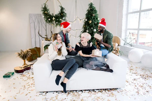 新年のお祝いで楽しい時を過すサンタ帽子で 人の若い友人のグループです 白いソファで 後ろにギフト ボックスと 人の男の子を保持している つの白人の女の子 Sitiing — ストック写真