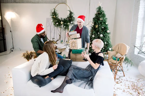 Group of happy people having a great time, celebrating New Year. Two girls and african boy sitting on white sofa and drinking champagne, and white boy behind is giving Christmas presents for them