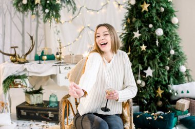 Genç ve güzel kız kot ve beyaz örgü kazak koltuk bir şekilde dekore edilmiş rahat oda Noel ağacı önünde oturuyor. Konfeti. Neşeli Noel, yeni yıl, Aile, tatil kavramı.