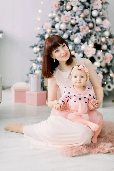 ピンクのドレスかわいい女の赤ちゃんと一緒に飾られたクリスマス ツリーの前の床の上に座って 楽しんで幸せな若い母親 メリー クリスマスとハッピー ホリデー — ストック写真