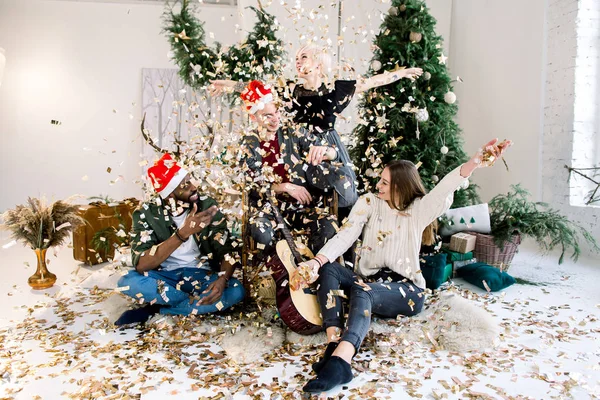 クリスマスと新年のパーティーのコンセプトです クリスマス ツリーと居心地の良い装飾が施されたリビング ルームでクリスマスを祝う 人の多民族の友人 紙吹雪フラッパー — ストック写真