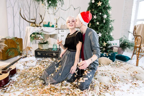 크리스마스 트리, 골든 색종이, 새 해, 행복 한 파티, 재미, 축 하를 던지고 근처 바닥에 앉아 사랑에 젊은 세련 된 부부 미소 — 스톡 사진