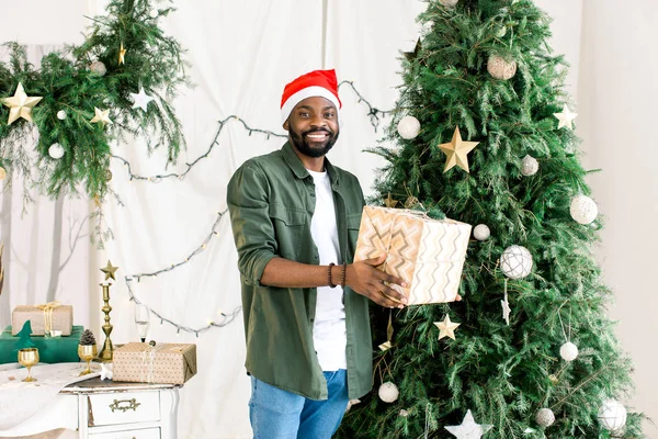 クリスマス ツリーの背景にギフト ボックスとサンタ帽子でスタイリッシュな緑のシャツの素晴らしい笑顔を着ているアフリカ系アメリカ人 — ストック写真