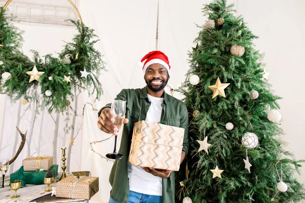 シャンパン ・ ギフト開催クリスマス ツリーの近くサンタ帽子で魅力的なアフリカ人を笑っています。ベンガルのライトです。居心地の良い客室です。クリスマス、新年、冬のコンセプト — ストック写真