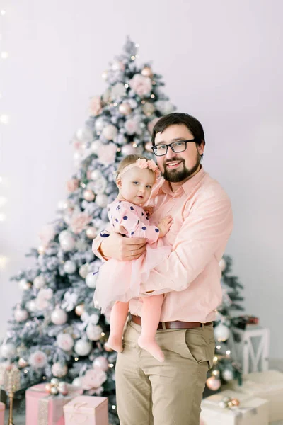 Feliz Natal. Jovem família celebrando o Natal em casa. Pai jovem em óculos segurando uma menina em adorável vestido de pêssego na frente da árvore de Natal — Fotografia de Stock