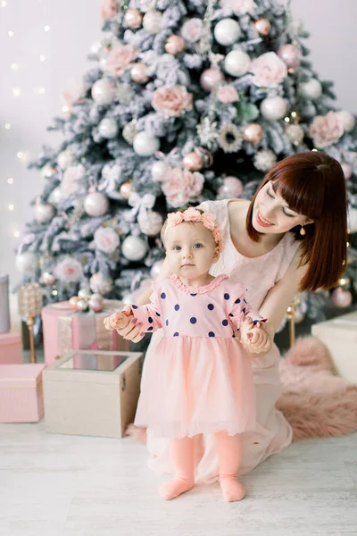 Jovem mãe em vestido rosa com seu pequeno um ano de idade daugter em vestido bonito se preparando para a árvore de Natal em casa . — Fotografia de Stock