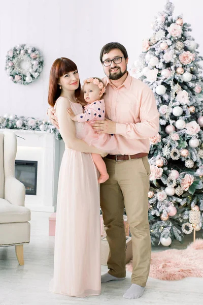 クリスマス ツリーの前に立って、カメラに笑顔のかわいい女の子との幸せな若い家族。冬、新年とクリスマスのコンセプトです。幸せな家族の概念 — ストック写真