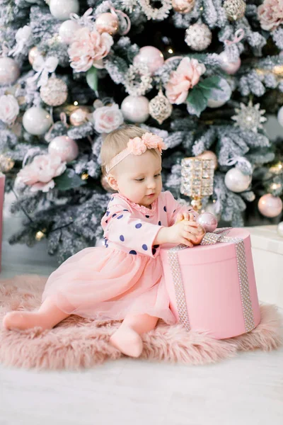 光のオレンジのドレスで幸せなかわいい女の子クリスマス プレゼントで遊ぶし、クリスマス ツリーの近くのピンクの毛皮のようなカーペットの上に座っています。家族、クリスマス、新年のコンセプト. — ストック写真