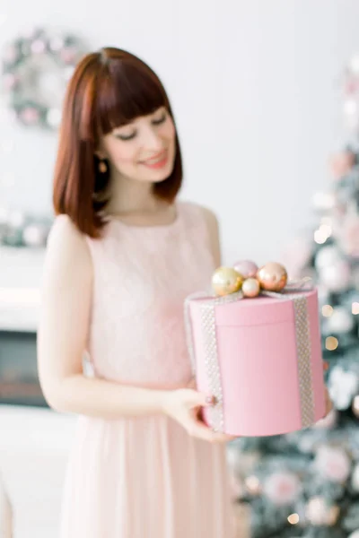 クリスマス プレゼント ボックスを保持している、クリスマスの装飾と美しい居心地の良いリビング ルームに立っている光のオレンジ色のピンクのドレスで、かなり若い女性を笑顔 — ストック写真