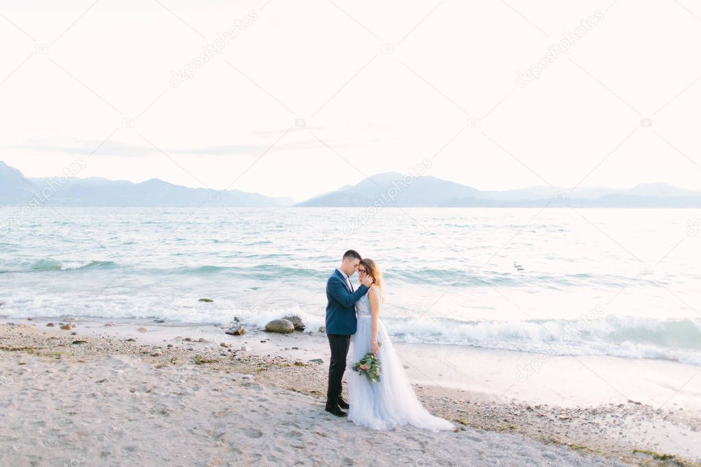 Tender portrait of young man hugging his pretty woman while standing on the pier near the lake on the wedding ceremony. Garda Lake, Italy