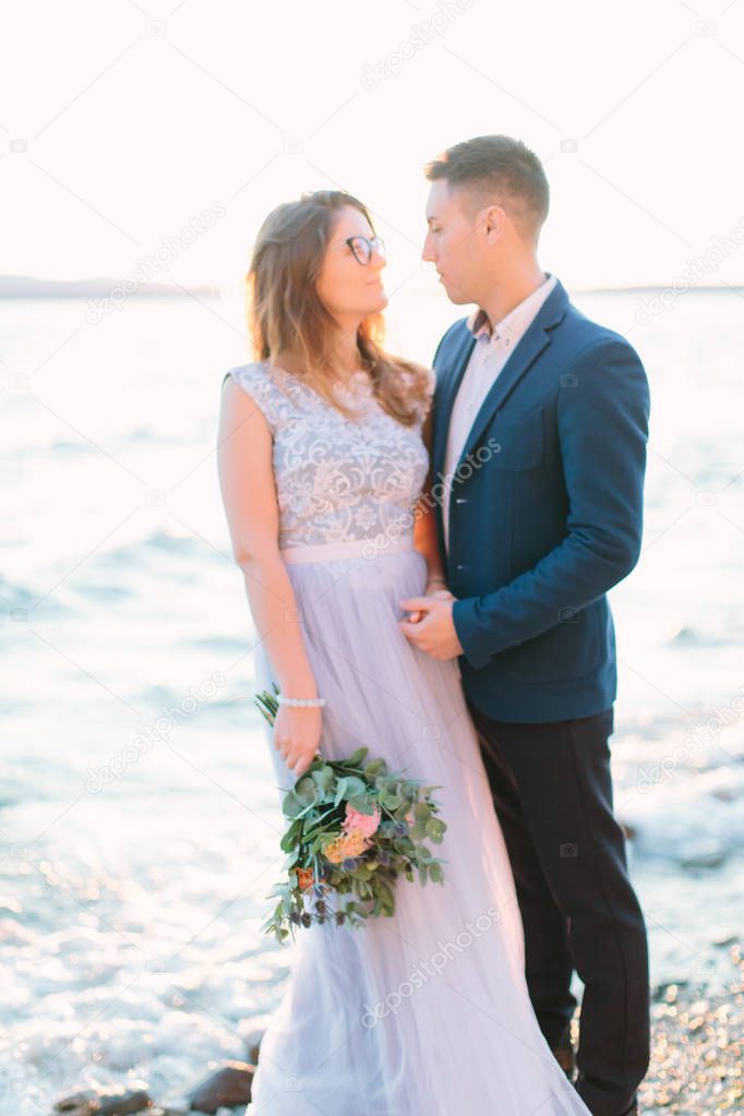 Happy groom and bride in beautiful wedding dress standing on the rocks near the Garda lake. Sirmione, Italy. Wedding couple