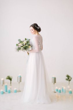 Beyaz duvarlı bir salonda poz ve zemin çiçek ve mum ile dekore edilmiş çiçek bir buket beyaz elbiseli güzel genç gelin.