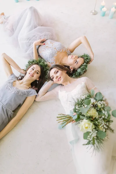 Top view of bride with female friends lying on the floor and smiling. Three young women lying on bed at home. Wedding morning before ceremony