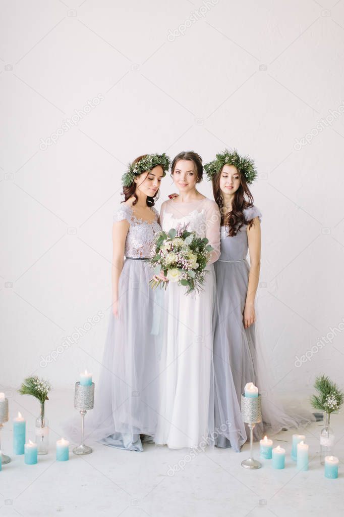 Happy bride in a white dress holds a wedding bouquet and poses with her friends in elegant dresses. White room is decorated with pine, flowers and blue candles.
