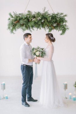 Mutlu gelin düğün buketi ve damat düğün töreninden sonra çam dekorasyon altında ayakta tutan beyaz elbiseli. Düğün töreni ışık beyaz odada çiçekler ve mumlarla süslenmiş.