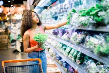 Pretty Afro young woman in jeans shopping organic veggies and fruits. Woman with trolley holding lettuce and radish in her arms clipart