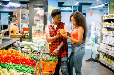 Young african couple in casual clothes choosing vegetables and fruits in grocery department of the supermarket, man is holding shopping basket clipart