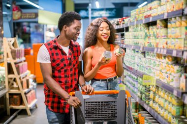 smiling african american couple with shopping trolley choosing food in supermarket clipart