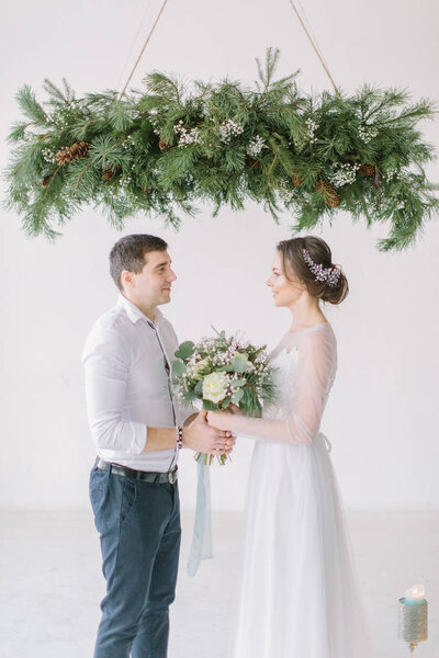 Happy bride in white dress holding wedding bouquet and groom standing under the pine decoration after wedding ceremony. Wedding ceremony in light white room decorated with flowers and candles.