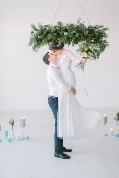 The groom lifted the bride up in his arms. The bride in luxury white wedding dress in the arms of the groom laughing. Wedding ceremony in light white room decorated with pine, flowers and candles.
