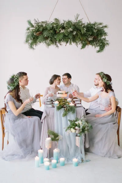 Group of people bride and groom, bridesmaids and groommen sitting at wedding table with wedding cake, pine decoration and candles in white decorated hall