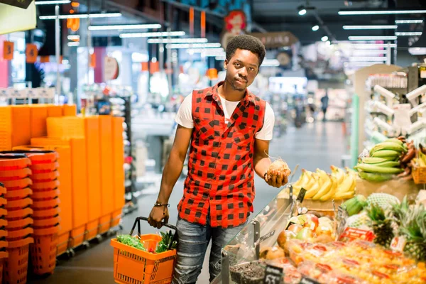 アフリカ系の若者は男のスーパー マーケットで食料品店で購入の青果です 黒人男性は 買い物籠を押しながら スーパー マーケットで野菜や果物を選択します スーパーで野菜を買い物男 — ストック写真