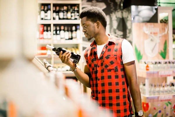 アフリカ人の人がスーパー マーケットでワイン セクションでのショッピングします ワインを買っている間市場で買い物をしている黒人の男性 ハンサムな男を保有する買い物かご製品の栄養値の読み取り — ストック写真