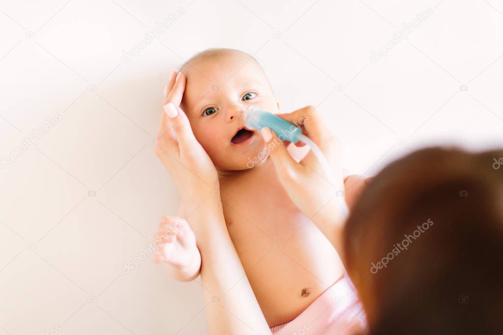 Mom holding her head to a small girl with her hands sucks the snot from the baby's nose lying on the bed through the aspirator. Cleaning the nose on the bed.