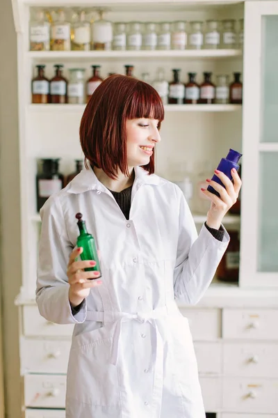 Smiling attractive young lady pharmacist holding green and blue glass bottles in her hands on the background of pharmacy interior