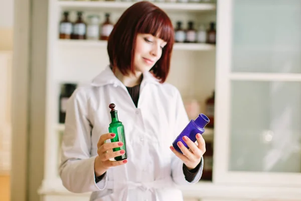 Smiling attractive young lady pharmacist holding green and blue glass bottles in her hands on the background of pharmacy interior