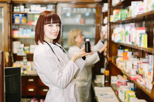 Smiling attractive young lady pharmacist holding a bottle of medicines in her hand with focus to her face. Other woman pharmacist choosing the drug on the background