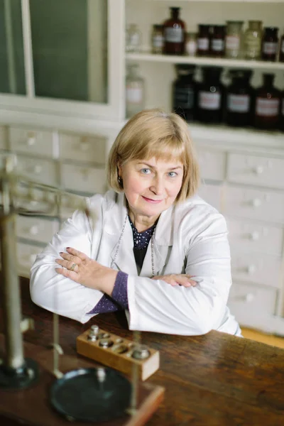 Smiling attractive middle-aged blond lady pharmacist sitting at the old wooden table with vintage scales in the pharmacy