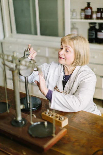 Smiling attractive middle-aged blond lady pharmacist holding her hands on the vintage scales in the laboratory room in pharmacy